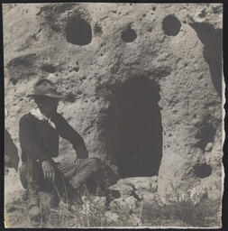 Man sitting beside doorway into cliff dwelling, Pajarito Plateau, New Mexico